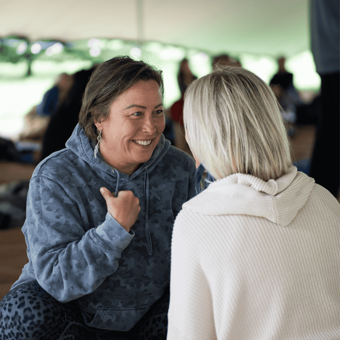 Two women engaged in a friendly conversation, smiling and making eye contact. The woman facing the camera is wearing a blue hoodie with a subtle pattern, while the woman with her back to the camera is wearing a cream-colored turtleneck. They are indoors, possibly at an event or gathering, with other people blurred in the background.