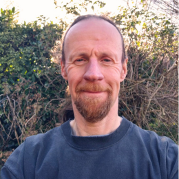 A man with a beard and receding hairline outdoors, smiling with trees and sunlight in the background.