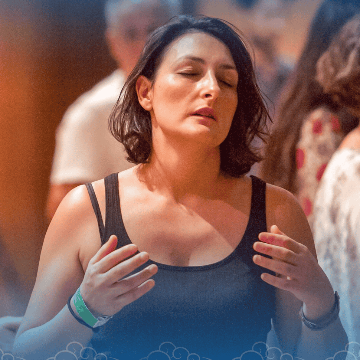 A woman with short dark hair and a black tank top appears to be praying or meditating with her eyes closed and hands over her chest in a crowded indoor setting.