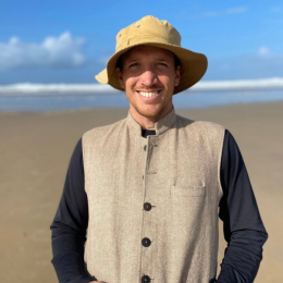 A man smiling at the beach, wearing a wide-brimmed hat and a beige vest over a black shirt.