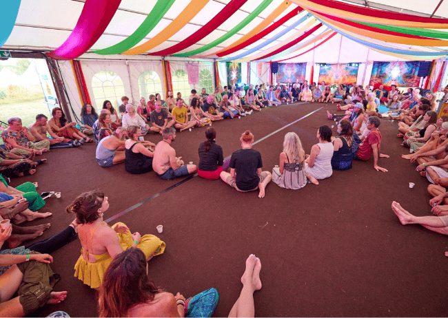 People sitting in a large circle inside a decorated tent with colorful ribbons hanging from the ceiling.