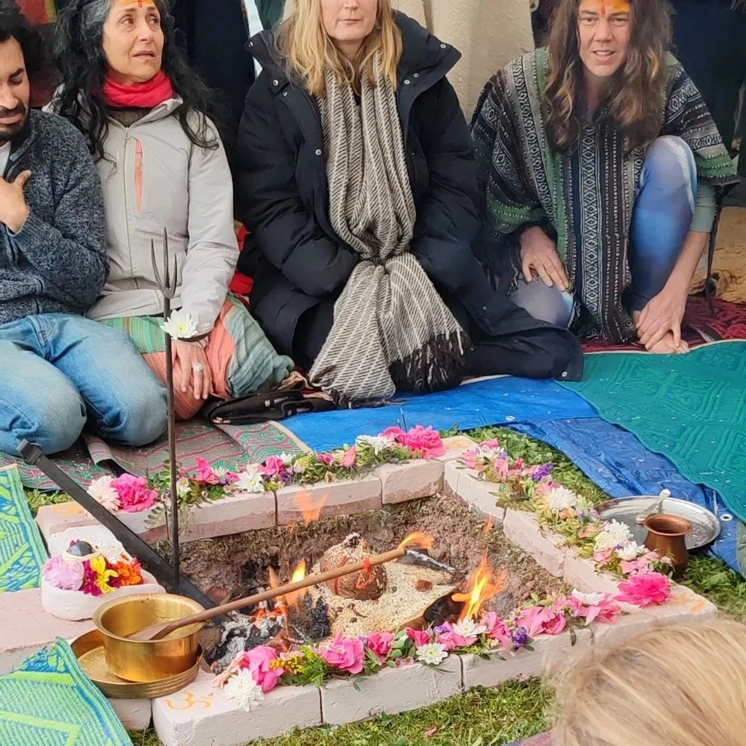 People participating in a spiritual or religious ceremony, sitting on mats around a small sacred fire decorated with pink and white flowers.