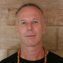 Portrait of a man with short blonde hair, blue eyes, wearing a black shirt and a wooden bead necklace, standing in front of a wooden wall.