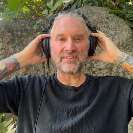 A smiling man with gray hair and beard wearing headphones outdoors, standing in front of a rock and green foliage.