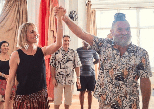 A group of people dancing and smiling in a brightly lit room with large windows, curtains, and hardwood floors.