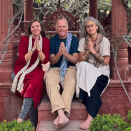 Three people sitting on steps praying with hands together, outdoors in a garden setting.