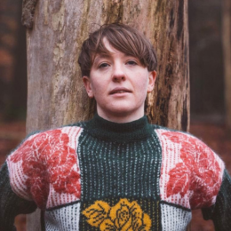 Person with short hair wearing a patterned sweater standing in front of a tree in an outdoor setting.