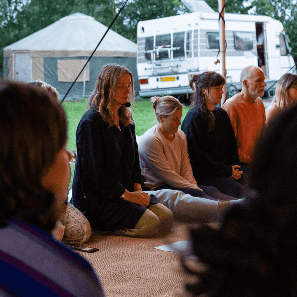 People sitting cross-legged on the ground with eyes closed, participating in a meditation or prayer session outdoors near a camping site with a caravan and tent visible in the background.
