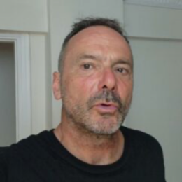 A middle-aged man with short dark hair and a beard, wearing a black shirt, standing indoors in front of a light-colored wall.