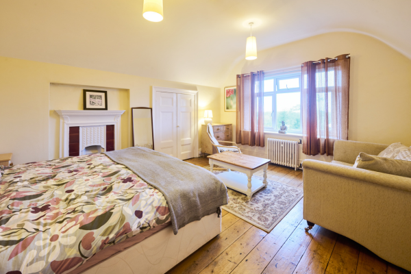 A cozy bedroom with a large bed, a decorative fireplace mantle, a white chair with a ottoman, a beige sofa, a window with curtains, and warm lighting.