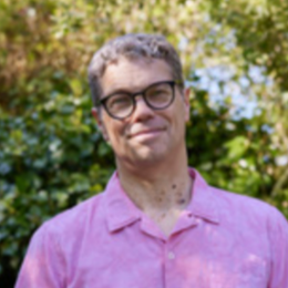 A man with curly hair, glasses, and a slight smile, wearing a pink polo shirt outdoors against a background of green trees and foliage.
