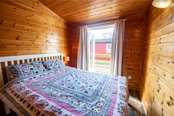 Bedroom with wooden paneled walls, a bed with floral quilt, white headboard, small lamp, open window with curtains, view of a red house outside.