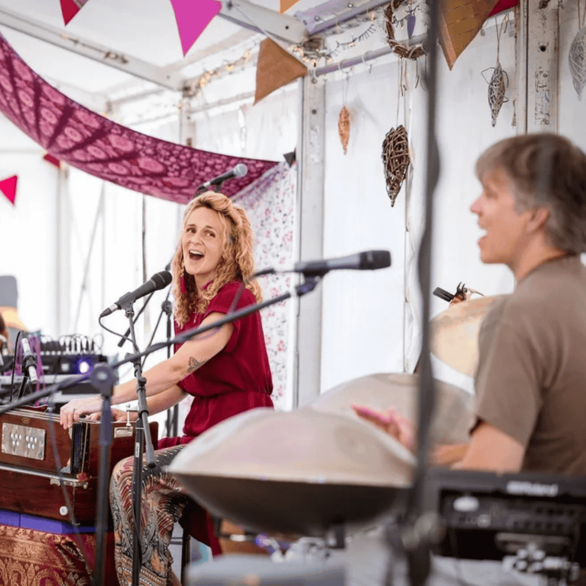 A woman with curly red hair singing into a microphone, playing a keyboard, and smiling, while another woman in a wheelchair is sitting nearby, smiling and clapping. The setting appears to be a decorated event space with bunting and hanging ornaments.