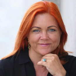 Portrait of a woman with red hair, wearing a black top and gold earrings, smiling with her hand resting on her chin.