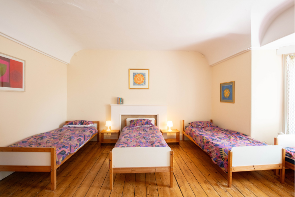 A room with three single beds with colorful patterned bedding, two nightstands with lamps, and framed artwork on the walls.
