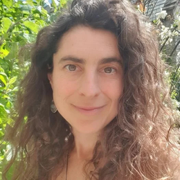 A woman with curly brown hair and fair skin smiling outdoors with greenery in the background.