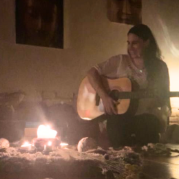 Person playing guitar and singing in front of candles in a cozy, dimly lit room.