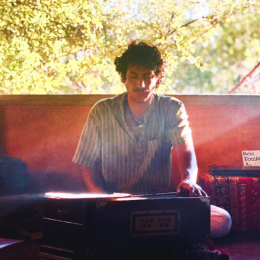 Young man seated on the ground, wearing a light-colored striped shirt, appears to be playing an electronic keyboard outdoors with autumn trees in the background.