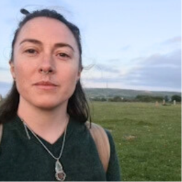 A woman with dark hair takes a selfie outdoors in a grassy field with a cloudy sky.