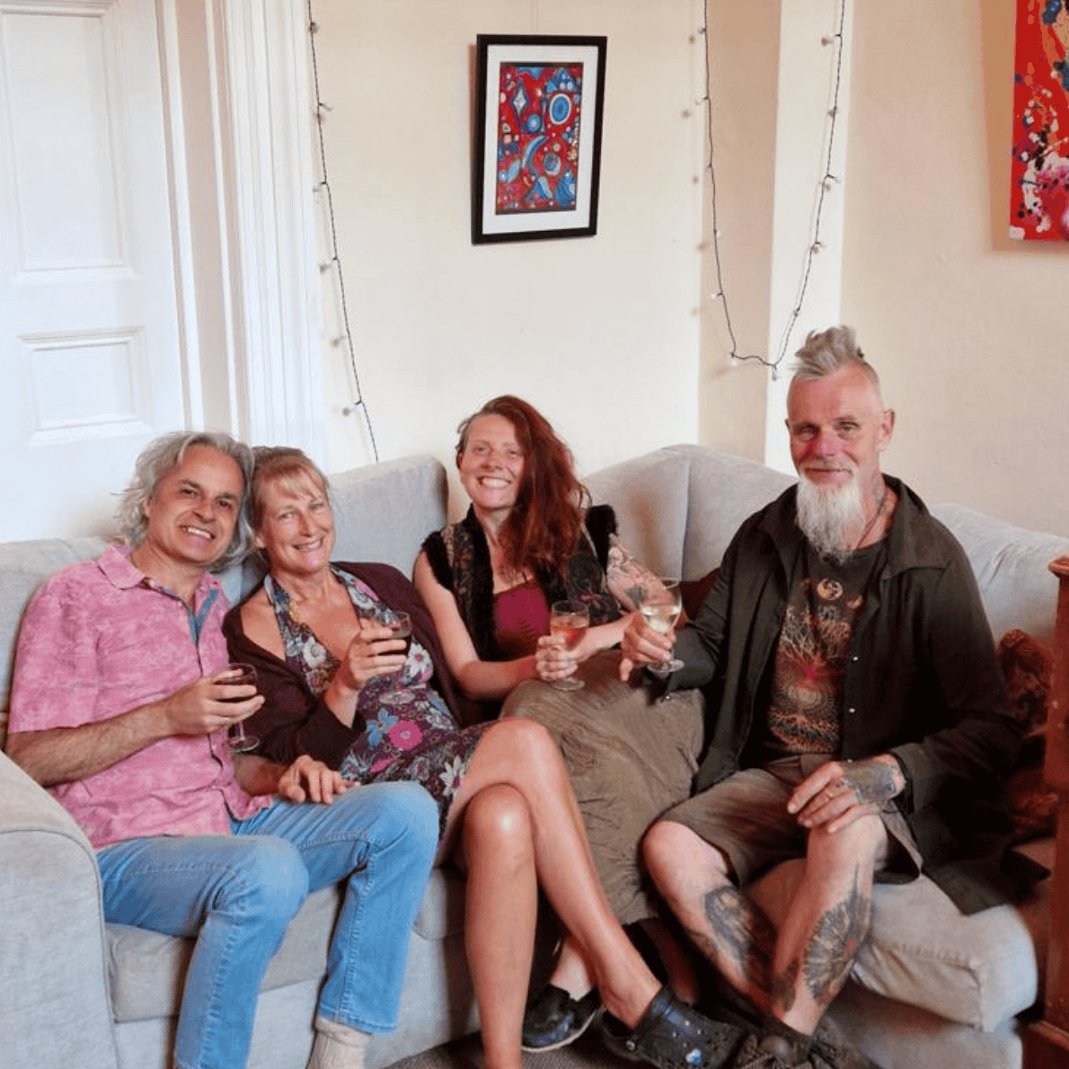Four people sitting on a beige couch holding drinks, smiling, and posing for the photo in a living room decorated with framed artwork and string lights.