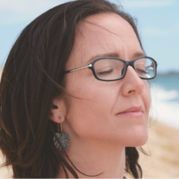 Close-up of a woman with dark hair and glasses, eyes closed, at the beach during daytime.