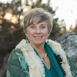 An elderly woman with short gray hair smiling outdoors, wearing a teal top and a green jacket with white fur lining.