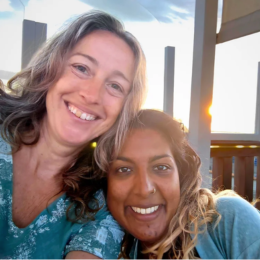 Two women smiling outdoors during sunset, one with blonde hair and the other with dark curly hair.