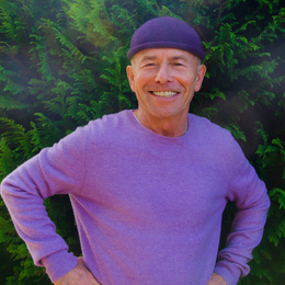 A smiling man wearing a purple sweater and dark cap standing outdoors in front of green foliage.