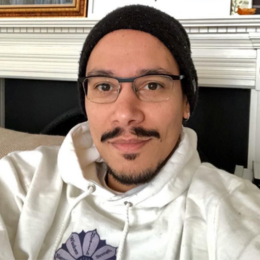 A man with glasses, a beard, and a mustache wearing a black beanie and white hoodie taking a selfie indoors.