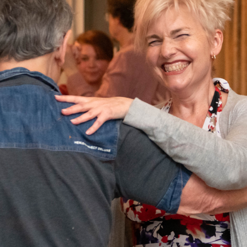 A woman with short blonde hair smiling and dancing with a man in a casual setting, with other people in the background.