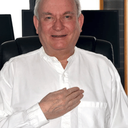 A man in a white shirt sitting in an office, making a gesture with his right hand over his chest, smiling.