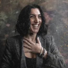 A woman smiling and touching her chest, wearing a blazer and bracelets, in front of a textured background.