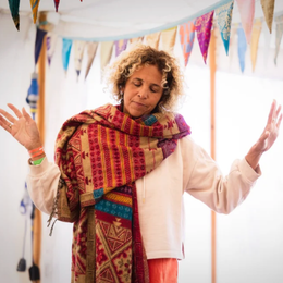 A woman standing with eyes closed and arms raised, wearing a colorful scarf and white jacket in a decorated indoor space.