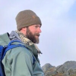 Profile of a man with a beard wearing a brown knit cap and outdoor gear, standing in a mountainous landscape.