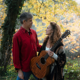 A man and woman stand facing each other outdoors in a park with autumn leaves, the woman holding a guitar.
