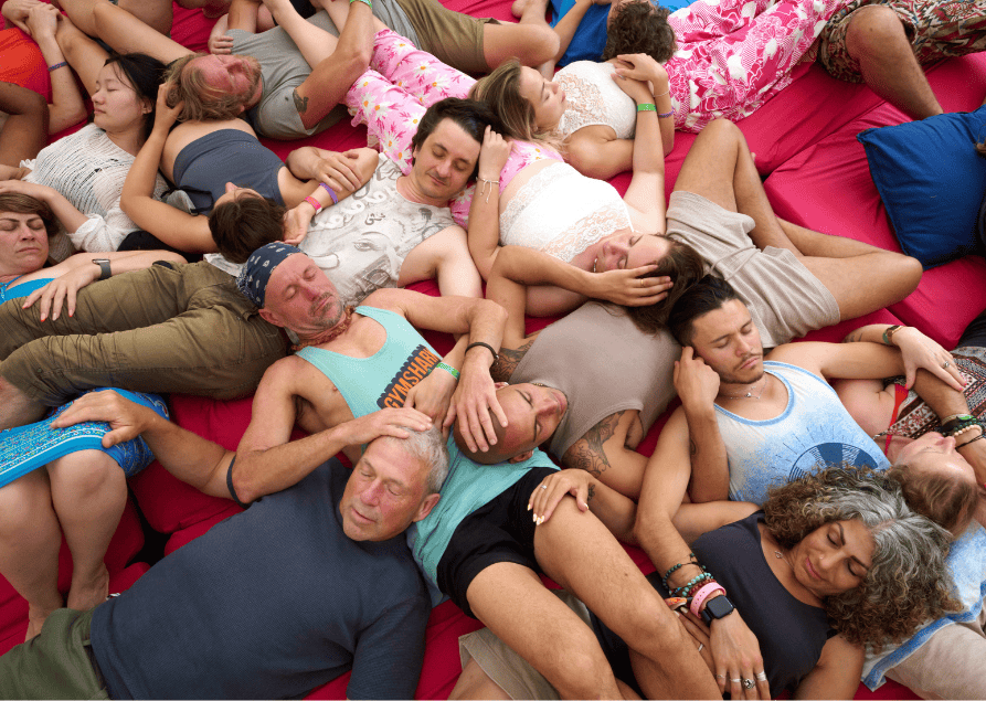 Multiple people of various ages sleeping on a large red mattress, some with pillows, in casual clothing.