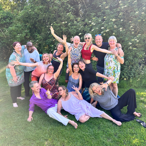 Group of twelve diverse adults outdoors on grass, posing happily with smile and playful gestures in front of green foliage.
