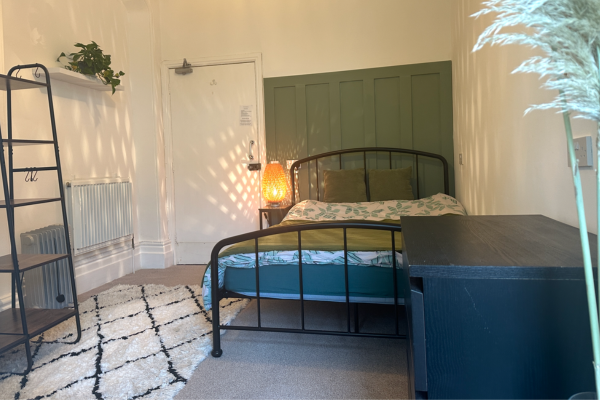 A bedroom with a black metal bed frame, green headboard cushions, a patterned duvet, a black dresser, a bedside lamp, a white and black rug, a white wall with a radiator, a black shelving unit, and a green door.