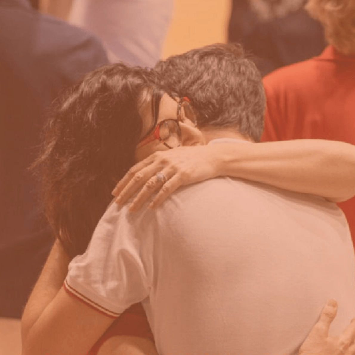 A couple hugging tightly, with the woman wearing glasses and the man having his back to the camera, in a warm, intimate setting.