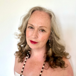 A woman with gray, curly hair and red lipstick, wearing a necklace with black and white beads, posing against a plain white background.