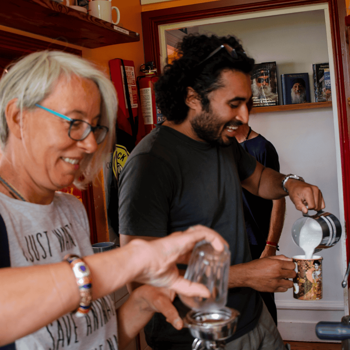 Two people, a woman with gray hair and glasses and a man with dark curly hair, smiling while pouring a beverage into a mug at a coffee shop.