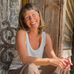 A woman with short brown hair smiling and laughing, wearing a white tank top, sitting outdoors with wooden and rustic elements in the background.