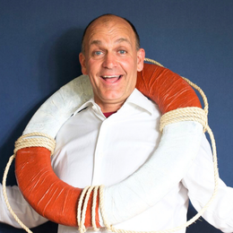 A man with a joyful expression wearing a white shirt, with a large lifebuoy draped around his shoulders, sitting against a dark blue background.