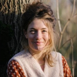 A woman with light brown hair smiling outdoors near a tree and tall grass.