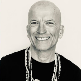 Black and white photo of a smiling man with a shaved head, wearing a black top and multiple beaded necklaces.