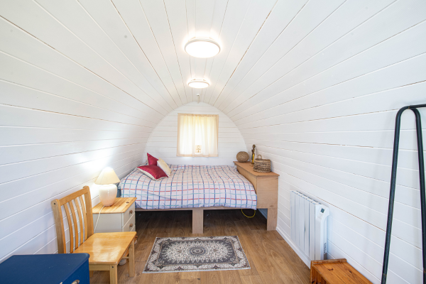 Small bedroom with white wooden panel walls, a bed with a checked blanket, a window with curtains, nightstand, lamp, chair, and small rug.