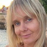 Close-up of a woman with blonde hair and blue eyes outdoors near a body of water.