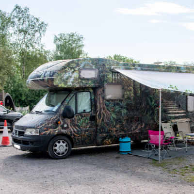 A camper van with a camouflage pattern parked outdoors with a pink chair and a blue barrel nearby, and a small orange traffic cone in front.