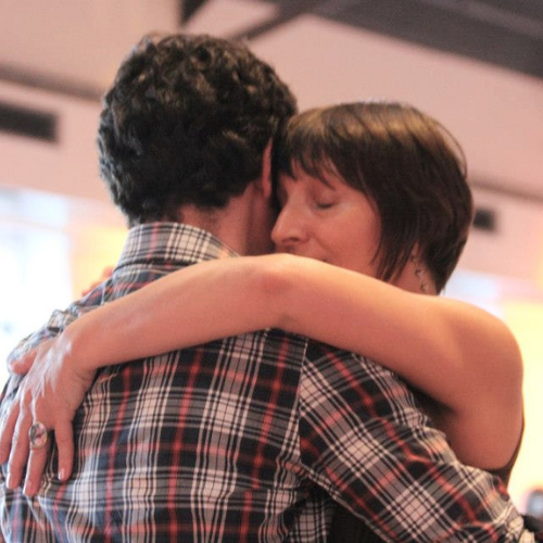 Two people hugging with their eyes closed in an indoor setting.