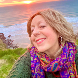 A woman taking a selfie outdoors near the ocean during sunset, wearing a colorful scarf and smiling.
